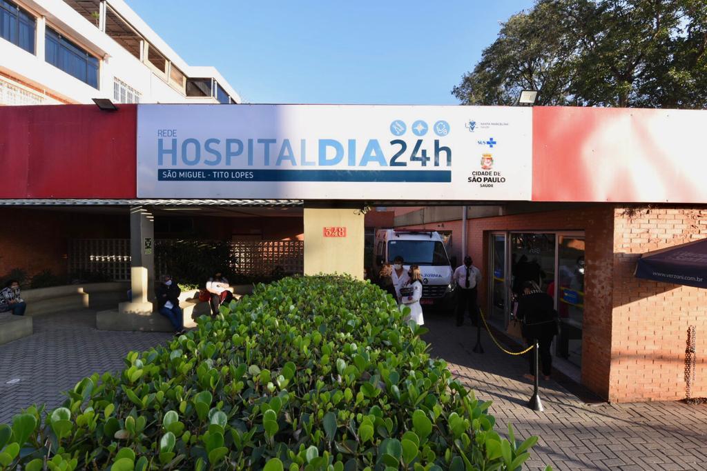 Foto de fachada. O prédio de paredes rosadas e faixa central vermelha exibe uma placa com Hospital Dia 24h e os logos do município, pessoas conversam diante do pátio que tem uma ambulância estacionada; à frente temos um detalhe de vegetação